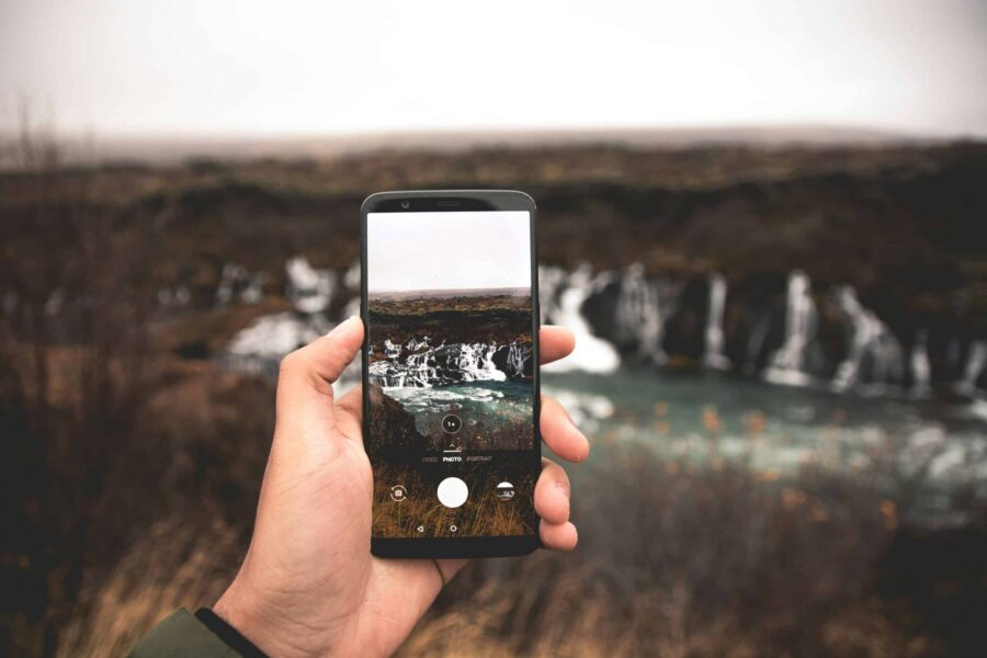 hand holding a cell phone and taking a photograph of a distant waterfall