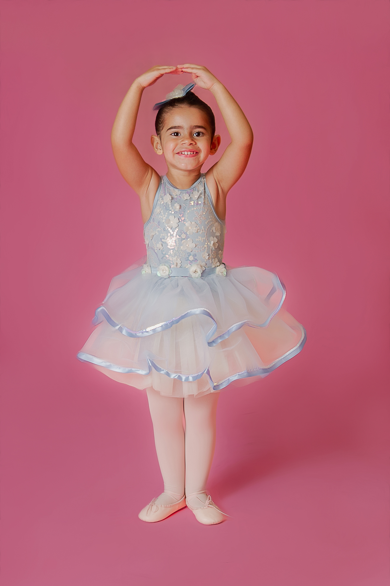 A young girl in a light blue and white tutu smiles with her arms raised above her head, posing against a pink background. She is wearing pink tights and ballet shoes, and a matching blue bow in her hair.