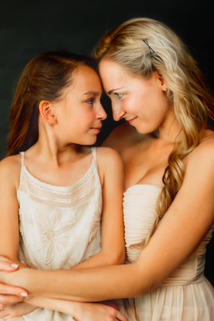 Studio portrait of Mom sitting with daughter in her lap and arms around her, facing each other with foreheads touching. Portrait by Jani Tzolov
