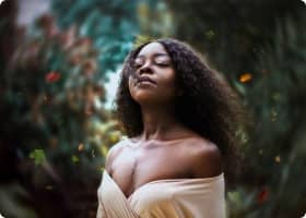 Black woman with eyes closed, wearing an off-the-shoulder beige top, amidst a blurred green foliage background
