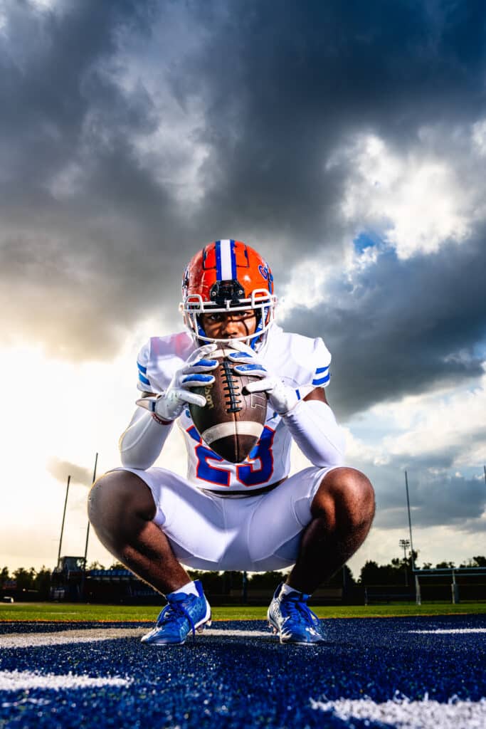 black teen boy in white football uniform posing on field with football in both hands
